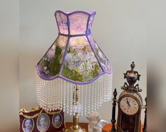 Puede incluir: Una pantalla de lámpara de color morado y verde con un patrón floral y flecos de cuentas blancas. La pantalla está sobre una base de lámpara dorada con un remate de porcelana blanca. La lámpara está sobre una mesa con un reloj y dos cuadros enmarcados.
