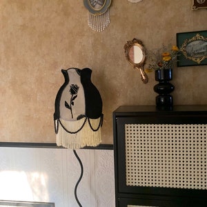 May include: A black and white lampshade with a black rose design and white fringe. The lampshade is on a black metal floor lamp with a curved stem. The lamp is in front of a black cabinet with a woven cane front.