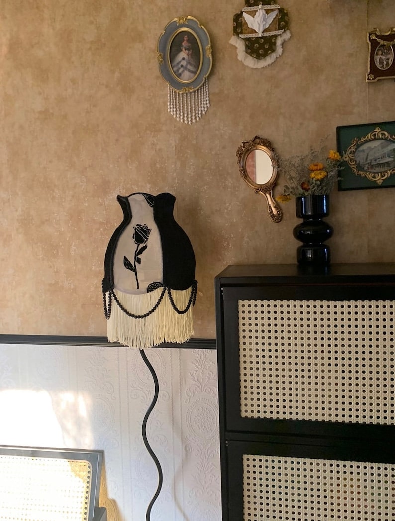 May include: A black and white lampshade with a black rose design and white fringe. The lampshade is on a black metal floor lamp with a curved stem. The lamp is in front of a black cabinet with a woven cane front.