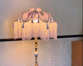 May include: A gold floor lamp with a white shade and a pink and purple floral design. The shade has a fringe trim and tassels.
