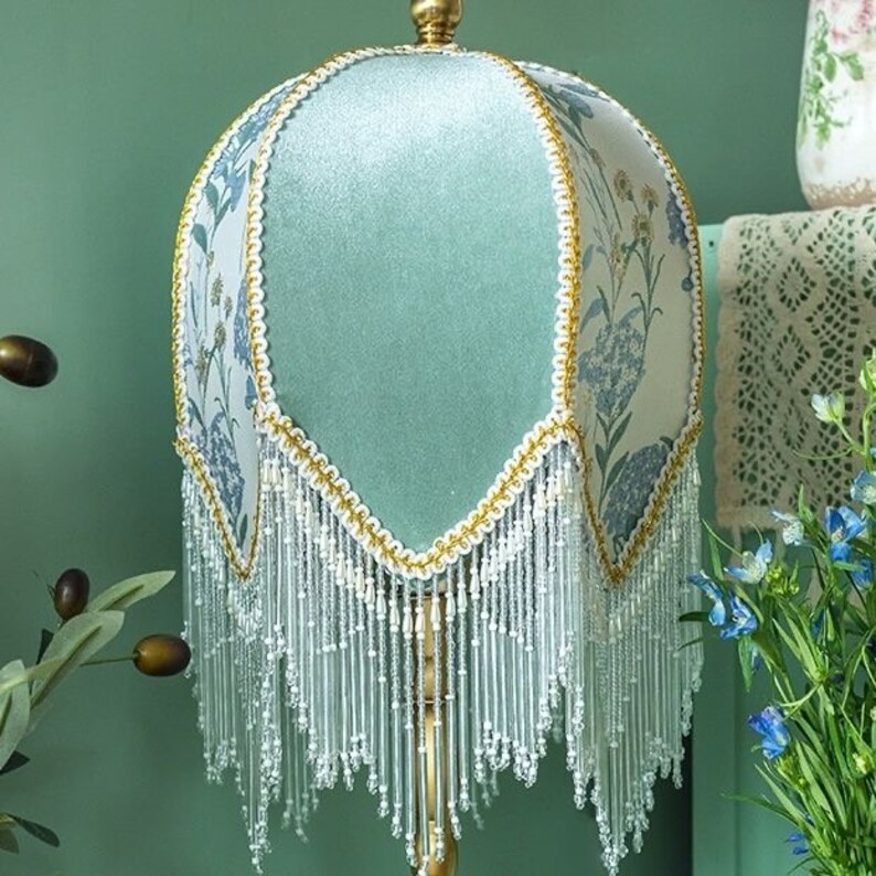 Puede incluir: Una pantalla de l&aacute;mpara azul claro con un patr&oacute;n floral y flecos blancos. La pantalla de la l&aacute;mpara est&aacute; hecha de tela y tiene un ribete dorado.
