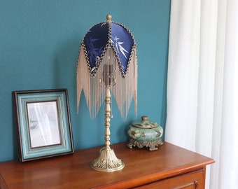 May include: A gold table lamp with a blue and white patterned lampshade with gold fringe. The lampshade has a geometric design with a floral pattern. The lamp is on a wooden table with a decorative ceramic jar.