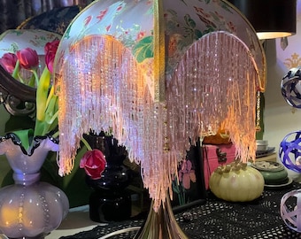 May include: A decorative table lamp with a floral patterned shade and beaded fringe. The lamp has a gold-coloured base and a white cord. The lamp is on a table with a black lace doily.