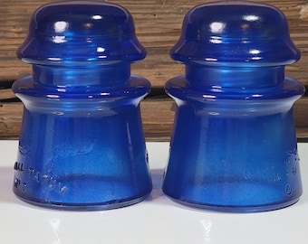 Peut inclure: Deux boutons isolants en verre bleu avec un sommet arrondi et une base large. Les boutons ont une finition brillante et irisée. Le texte "MADE IN U.S.A." est visible sur un bouton.