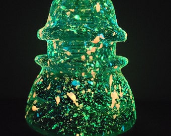 Op de afbeelding: Een groene glazen isolator met witte vlekken, die gloeit onder een blacklight. De isolator heeft een afgeronde bovenkant en een brede basis.