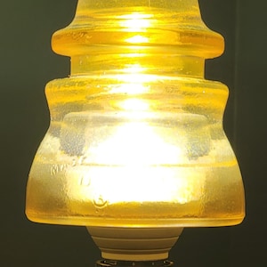 May include: A vintage, yellow glass insulator with a layered design. The glass has a slight texture and is mounted on a black and white base. The insulator is illuminated from within, creating a warm glow.