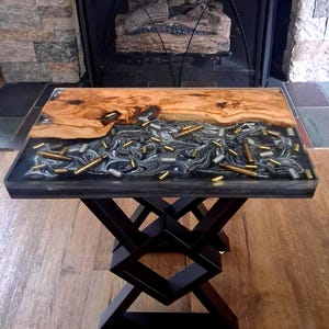 May include: A rectangular side table featuring a live edge wood slab joined with black and white marbled resin. The resin section is embedded with numerous metallic bullet casings in brass and silver tones. The table stands on a black metal base.