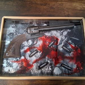 May include: A framed artwork featuring a detailed depiction of a revolver with a brown handle, set against a backdrop of red and black splatters and scattered bullets. The artwork is encased in a gold-colored frame.