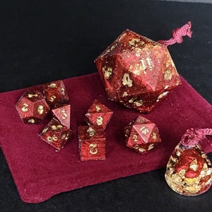 May include: A set of polyhedral dice in various shapes, including a large d20, with a rich red hue and gold leaf accents. The dice are displayed on a burgundy velvet pouch, suggesting a gaming theme.