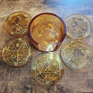 May include: Set of five resin coasters with a matching holder. The coasters are round, with gold geometric designs and numbers, and a brown and gold color scheme. The holder is a matching brown and gold color.