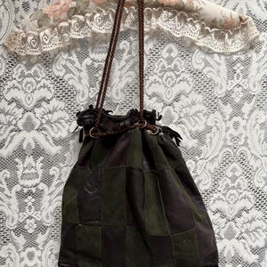 Könnte beinhalten: Ein Beutel mit Kordelzug aus einem Patchwork aus dunkelbraunen und olivgrünen Lederquadraten. Die Tasche verfügt über zwei dünne, geflochtene braune Kordelriemen und eine Reihe dunkelbrauner Lederfransen entlang der Unterkante des Hauptteils.