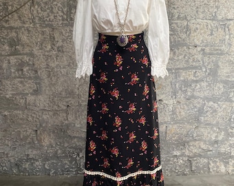 Pode incluir: Uma saia maxi preta com estampa floral e acabamento em renda branca. A saia é usada com uma blusa branca e um colar de corrente prateada com um pingente de pedra roxa.