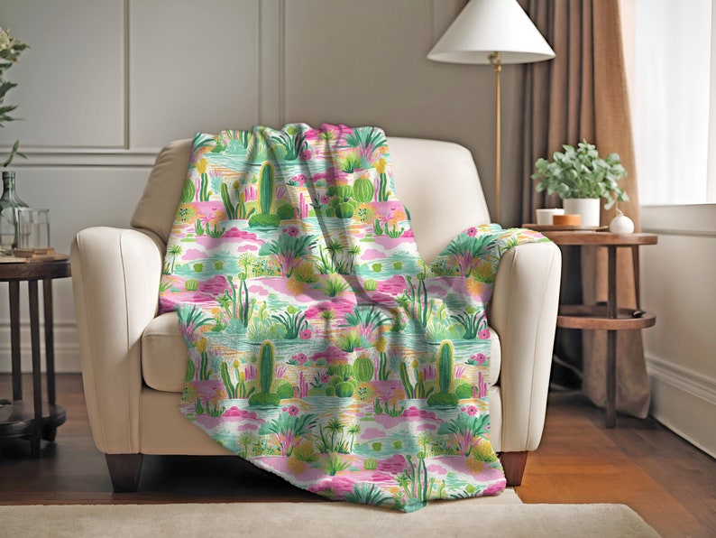 Puede incluir: Una manta de tiro rosa, verde y amarilla con un patr&oacute;n de paisaje de cactus y desierto. La manta est&aacute; drapeada sobre un sill&oacute;n blanco.