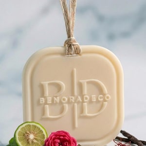 May include: Cream-colored, square-shaped air freshener with rounded corners, featuring the embossed text "BENORADECO". A natural twine loop is attached. The image includes a lime slice, a pink rose, and spices, suggesting a fragrance theme.