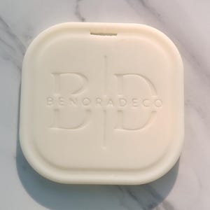 May include: Off-white, square-shaped decorative item with rounded edges. The item has a recessed design with the letters "BD" and the text "BENORADECO" embossed on the surface. A small notch is visible at the top edge.