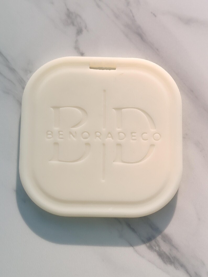 May include: Off-white, square-shaped decorative item with rounded edges. The item has a recessed design with the letters "BD" and the text "BENORADECO" embossed on the surface. A small notch is visible at the top edge.