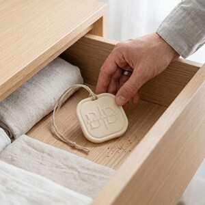 May include: A square, off-white scented sachet with the embossed text "MEMORADECO" is held above an open wooden drawer. The drawer contains folded linens in neutral tones. A natural twine loop is attached to the sachet.