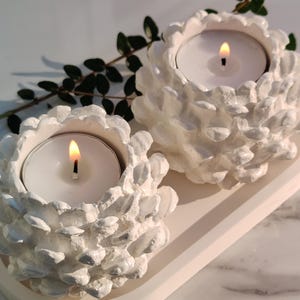 May include: Two white ceramic candle holders shaped like pine cones with tea lights inside. The candle holders are on a white tray.