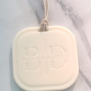 May include: Cream-colored, square-shaped decorative item with rounded edges, suspended by a natural twine loop. The embossed text reads "BENORADECO" with stylized initials. The item has a smooth, matte finish.