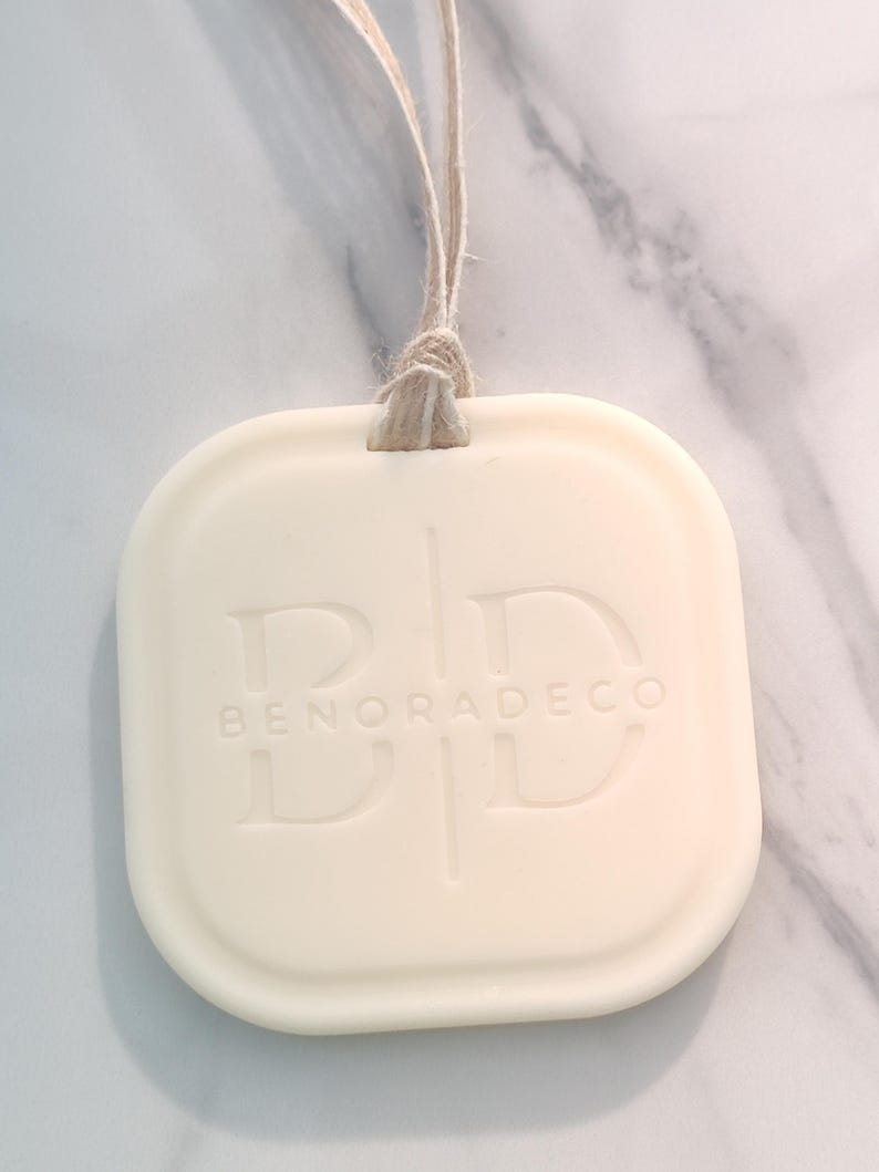 May include: Cream-colored, square-shaped decorative item with rounded edges, suspended by a natural twine loop. The embossed text reads "BENORADECO" with stylized initials. The item has a smooth, matte finish.