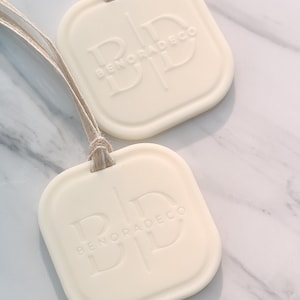 May include: Two off-white, square-shaped air fresheners with rounded corners and the embossed text "BENORADECO". Each freshener is attached to a natural-colored string. The background is a white and gray marble pattern.