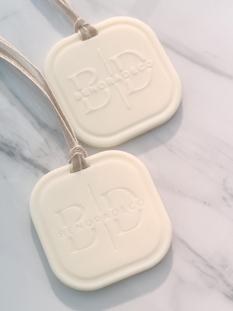 May include: Two off-white, square-shaped air fresheners with rounded corners and the embossed text "BENORADECO". Each freshener is attached to a natural-colored string. The background is a white and gray marble pattern.