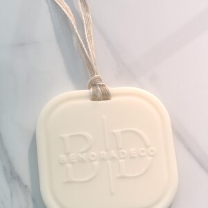 May include: Off-white, square-shaped air freshener with rounded corners and a beige string for hanging. The embossed text reads "BENORADECO" with stylized initials. The background is a marble-patterned surface.