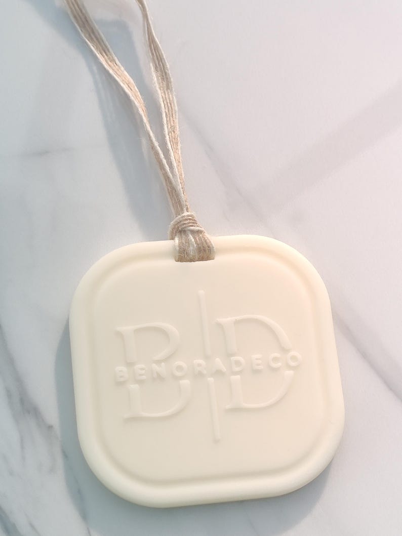 May include: Off-white, square-shaped air freshener with rounded corners and a beige string for hanging. The embossed text reads "BENORADECO" with stylized initials. The background is a marble-patterned surface.