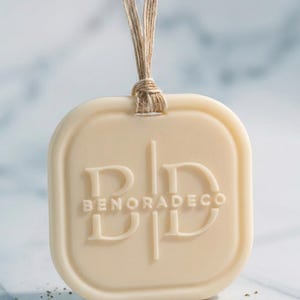 May include: A cream-colored, rectangular air freshener with rounded corners, suspended by a natural twine cord. The embossed text "BENORADECO" is centered on the front. The background is a light, blurred surface.