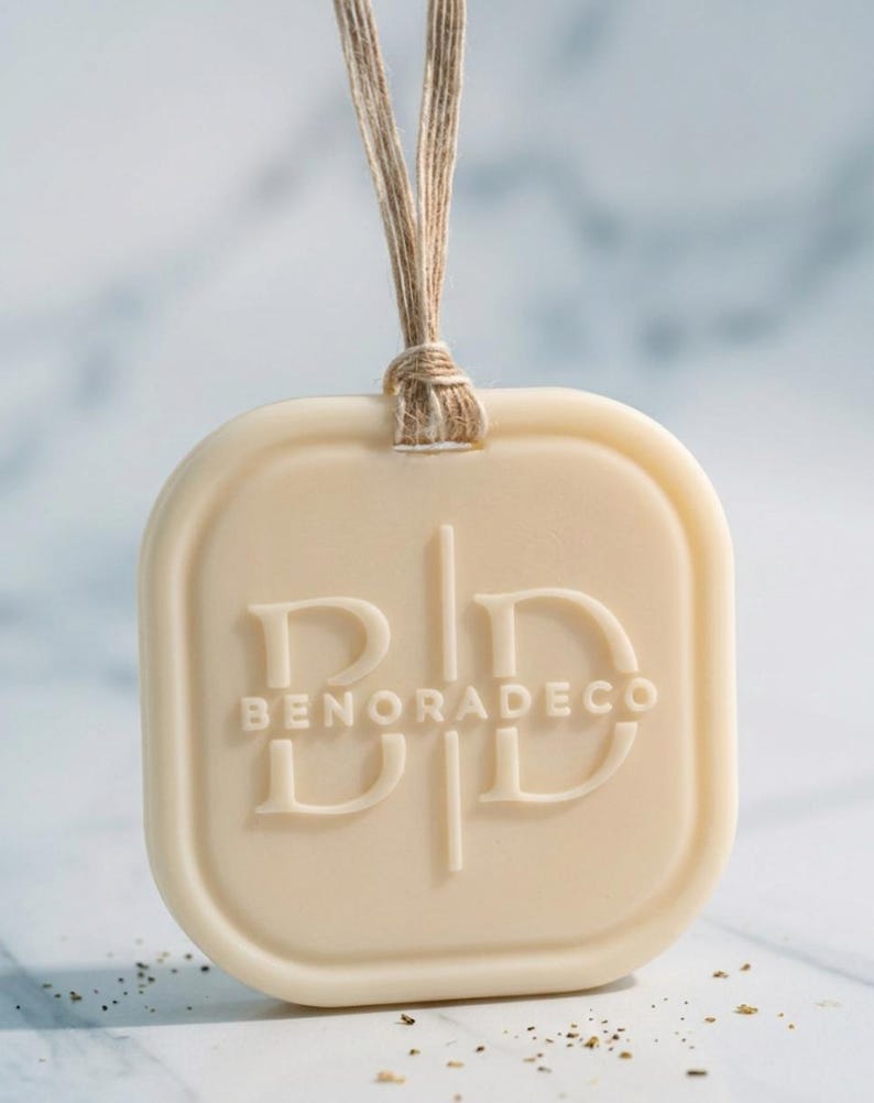 May include: A cream-colored, rectangular air freshener with rounded corners, suspended by a natural twine cord. The embossed text "BENORADECO" is centered on the front. The background is a light, blurred surface.