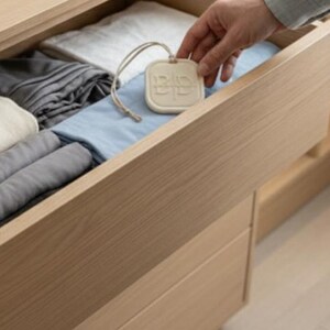 May include: A wooden drawer filled with neatly folded linens in shades of white, gray, and blue. A hand holds a cream-colored, rectangular tag with a string, likely a scented sachet or drawer freshener, adding a touch of fragrance to the storage space.