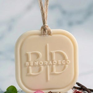 May include: Cream-colored, rectangular air freshener with rounded corners, embossed with the brand name "BENORADECO". A natural twine loop is attached. The image includes rose petals, spices, and herbs, suggesting a natural fragrance.
