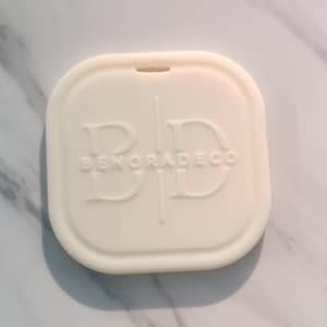 May include: Off-white, square-shaped decorative item with rounded edges. The item has the text "BENORADECO" embossed on the surface. The item is placed on a white and gray marble surface.