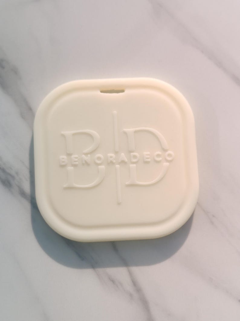May include: Off-white, square-shaped decorative item with rounded edges. The item has the text "BENORADECO" embossed on the surface. The item is placed on a white and gray marble surface.