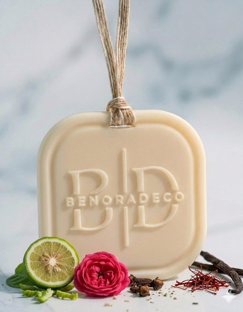 May include: Cream-colored, square-shaped air freshener with rounded corners, featuring the embossed text "BENORADECO". A natural twine loop is attached. The image includes a lime slice, a pink rose, and spices, suggesting a fragrance theme.
