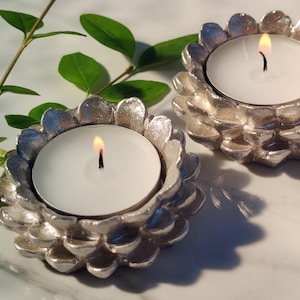 May include: Two silver-colored, flower-shaped candle holders with lit white candles. The candle holders have a textured petal design. Green leaves are visible in the background, adding a natural element to the scene. The candles are burning with a warm, inviting glow.