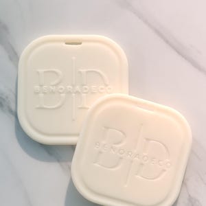 May include: Two off-white, square-shaped soap dishes with rounded corners. Each dish has the embossed text "BD BENORADECO" and a small notch at the top. The dishes are arranged on a marble surface.