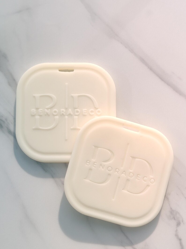 May include: Two off-white, square-shaped soap dishes with rounded corners. Each dish has the embossed text "BD BENORADECO" and a small notch at the top. The dishes are arranged on a marble surface.