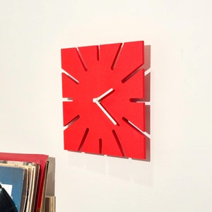 May include: A red clock with white hands and a unique design. The clock has a square shape with a sunburst pattern.