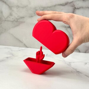 May include: A red heart-shaped box with a middle finger sticking out of the top. The box is open and the finger is visible.