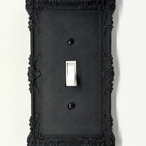 Puede incluir: Cubierta de interruptor de luz negra con un diseño ornamentado y enmarcado. La cubierta rectangular presenta un interruptor basculante y dos tornillos. El marco tiene patrones florales y de volutas detallados. El interruptor es blanco.