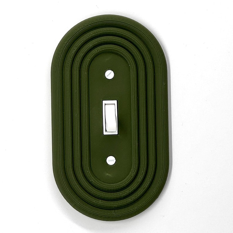 Op de afbeelding: Een olijfgroene lichtschakelaar cover met een decoratief, gelaagd ovaal ontwerp. Het heeft een witte tuimelschakelaar en twee kleine schroeven. Het ontwerp is een moderne, minimalistische stijl.