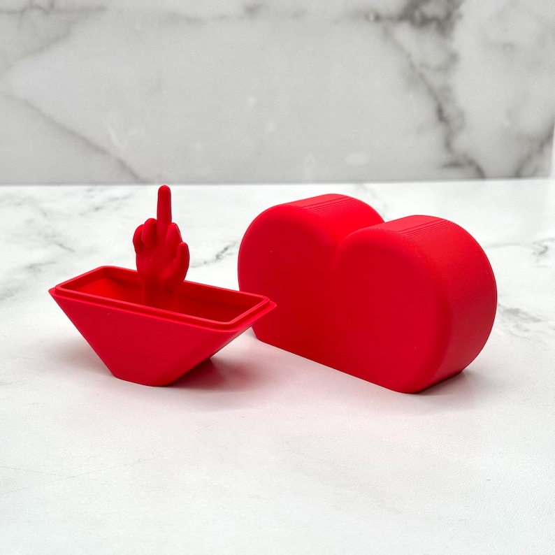 May include: A red heart-shaped box with a lid that opens to reveal a small red hand giving the middle finger.