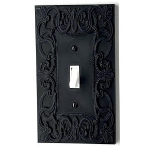 May include: Black decorative light switch cover with an ornate floral and scrollwork design. The rectangular cover has a single toggle switch and two screws. The switch is white, and the cover is mounted on a white wall.