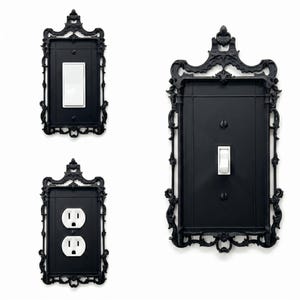 May include: Three black wall plates with ornate, decorative frames. One plate features a white rocker switch, one features a white duplex electrical outlet, and the largest plate on the right features a white toggle switch with two black screws.