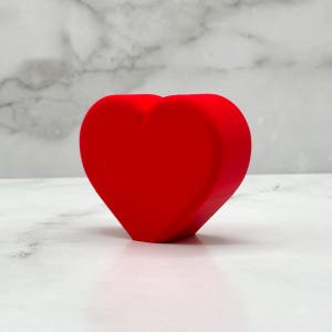 May include: A bright red heart-shaped object made of a smooth, solid material. The heart is sitting on a white marble surface.
