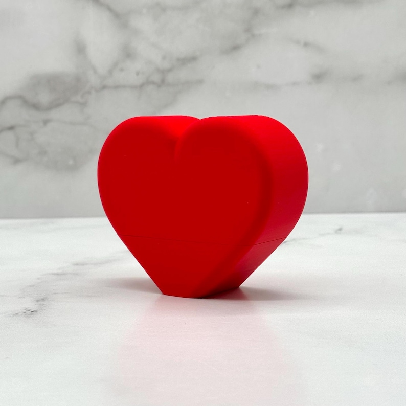 May include: A bright red heart-shaped object made of a smooth, solid material. The heart is sitting on a white marble surface.