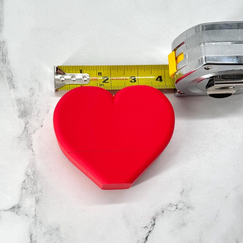 May include: A bright red heart-shaped object is measured with a yellow tape measure. The tape measure reads 4 inches.