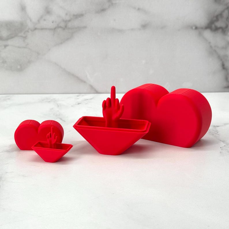 May include: Three red heart-shaped boxes, one larger than the other two. The largest box is open and the middle box is partially open. The smallest box is closed. The middle box has a small red hand with a middle finger raised inside.
