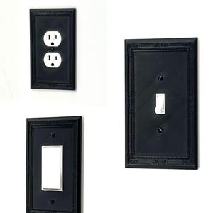 May include: Three black electrical outlet and light switch covers with ornate, framed designs. The covers include a two-outlet receptacle, a single rocker switch, and a single toggle switch. The white outlets and switches contrast with the black frames.